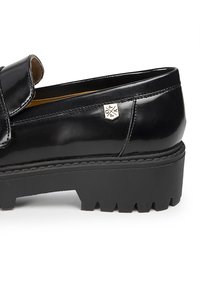 Zapato de charol negro con una correa ancha, suela de goma negra gruesa y un pequeño logotipo cerca del talón. Textura suave y diseño elegante.