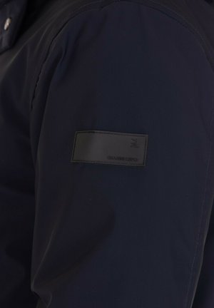 Manica di giacca blu scuro con una patch rettangolare nera che presenta le iniziali in rilievo "GL" e il nome del marchio "GIANNI LUPO".