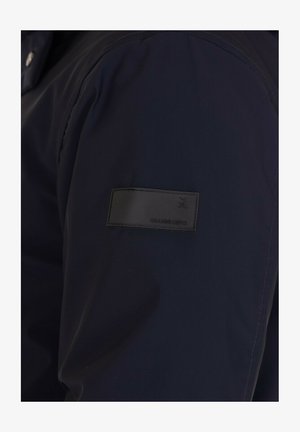 Manica di giacca blu scuro con una patch rettangolare nera che presenta le iniziali in rilievo "GL" e il nome del marchio "GIANNI LUPO".