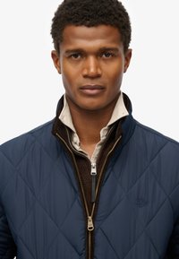 Man met kort krullend haar, gekleed in een beige shirt met een bruine gebreide ritsjack en een marineblauw gewatteerd jack, die recht vooruit kijkt.