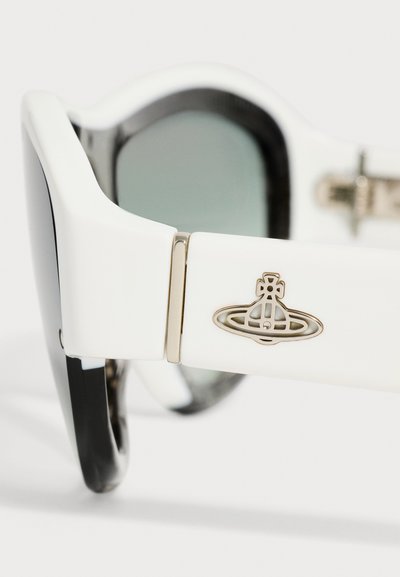 Branches de lunettes de soleil blanches et noires présentant un logo argenté en forme de planète et une charnière métallique avec des verres teintés en arrière-plan.