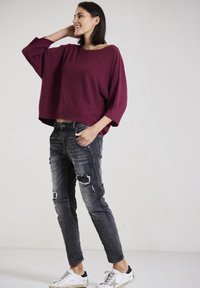Burgunderfarbener Oversized-Pullover mit weiten Ärmeln, kombiniert mit ausgebleichten schwarzen Jeans. Weiße Sneaker mit schwarzen Akzenten runden den Look ab.