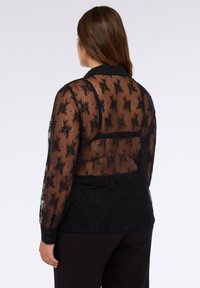 Blusa nera trasparente con motivo in pizzo floreale, maniche lunghe e colletto. Tessuto morbido con una parte inferiore aderente.
