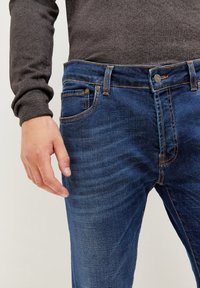 Jeans i mörkblå denim med en subtil blekt textur, som har kontrasterande orange sömmar och en femficksdesign, bärs med en grå tröja.