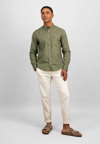 Camicia a maniche lunghe verde oliva con colletto a camicia, abbinata a pantaloni bianchi a sigaretta e sandali marroni. La camicia presenta una tasca sul petto.
