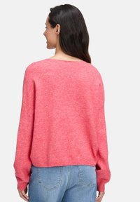 Frau mit langen dunklen Haaren, die einen lockeren rosa Pullover und hellblaue Jeans trägt, von hinten vor einem schlichten weißen Hintergrund.