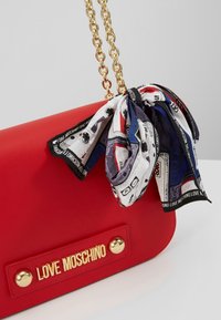 Punainen nahkalaukku, jossa on ketjuhihna, kultaisen sävyinen logolevy ja koristeellinen silkkihuivisolmu sinivalkoisena, jossa on kuvioituja painatuksia.