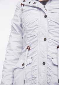 Veste blanche avec côtés froncés, boutons pression et cordons de serrage. Dotée de deux poches avant et d'une surface texturée.