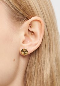Nærbillede af et blond øre med to guld øreringe: en lille stud i øreflippen og en buet barbell i det indre ørebrusk.