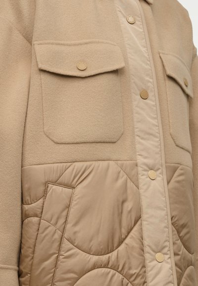 Μπεζ εξωτερικό μπουφάν με καπιτονέ κάτω μέρος, δύο τσέπες στο στήθος, μεταλλικά κουμπιά πατιλέτας και απαλό, υφασμάτινο φινίρισμα με υφή.