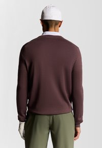 Maglione marrone con texture, maniche lunghe, colletto girocollo e orlo leggermente più corto. Abbinato a pantaloni verde oliva e un cappellino bianco. Guanti sulla mano sinistra.