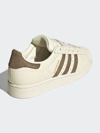 Biela kožená teniska s béžovými semišovými detailmi a tromi hnedými pruhmi. Má gumovú podrážku a na boku nápis „SUPERSTAR“.