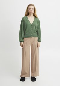 Zelený pletený cardigan s výstřihem do V a pěti knoflíky, v kombinaci s širokými béžovými kalhotami a černými platformovými botami. Hladká textura, uvolněný střih.