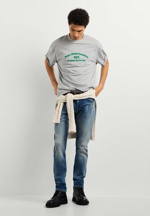 Uomo che indossa una t-shirt grigia Pepe Jeans London, jeans blu, stivali neri e un maglione beige chiaro legato in vita, guarda verso il basso.