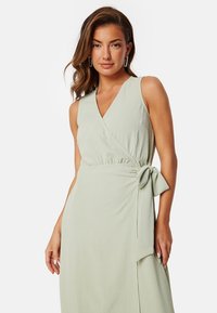 Světlezelené zavinovací šaty vyrobené z jemné tkaniny; mají výstřih do V, bez rukávů a vázání v pase.