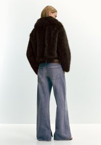 Osoba se světlými hnědými vlasy, oblékající tmavě hnědou umělou kožešinovou bundu a široké modré džíny, stojící na pozadí prosté bílé zdi.