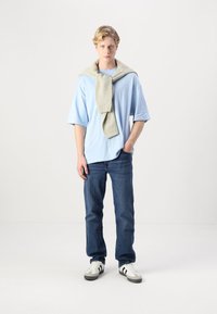 Light blue short-sleeve shirt, beige knitted sweater draped over shoulders, blue jeans, and white sneakers with black stripes.