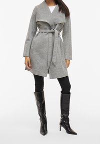 Szary płaszcz z szerokim kołnierzem, z paskiem, wykonany z teksturowanej tkaniny. Połączony z czarnymi, sięgającymi do kolan, spiczastymi botkami i czarnymi legginsami.