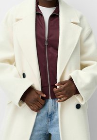 Witte wollen jas met een ontspannen pasvorm, grote revers en zwarte knopen, gedragen over een bordeauxrode jas met rits en lichtblauwe spijkerbroek.