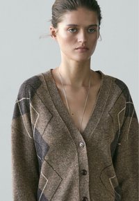 Młoda kobieta z zaczesanymi do tyłu włosami, ubrana w brązowy i szary sweter na guziki w romby oraz cienki srebrny naszyjnik na neutralnym tle.