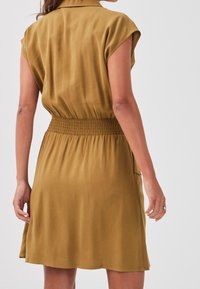 Robe à manches courtes de couleur moutarde avec une taille froncée. Tissu lisse, détail de col et poches latérales visibles.