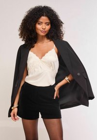 Sieviete ar čirkainiem matiem, tērpusies melnā žaketē, kas piesegta pār pleciem, krēmkrāsas mežģīņu apmalotā topā, melnās biksēs un zeltainos rokassprādzēs, pozē pārliecinoši.