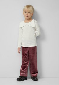 Junges Mädchen mit blonden Haaren, das ein weißes Langarmshirt mit Rüschensaum, lila Samthose mit kleinen Punkten und schwarze Schuhe trägt und steht.