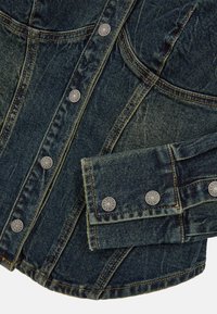Donkerblauwe spijkerjack met zilveren drukknoopjes, zichtbare stiksels en schuine naden. Beschikt over een omgeslagen manchet met een rafelige rand.