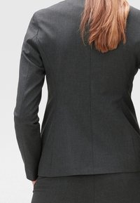 Blazer grigio su misura, tessuto testurizzato, silhouette strutturata, con uno spacco singolo sul retro, maniche adattate, con cuciture sottili e senza embellishments visibili.
