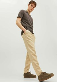 Polo shirt grigio scuro, pantaloni beige e scarpe con lacci marroni. L'outfit presenta un design aderente, tasche visibili e materiali testurizzati.
