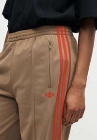 Pantalon de survêtement beige avec des bandes orange et un logo orange, doté d'une poche zippée sur le côté droit, porté avec une chemise noire.