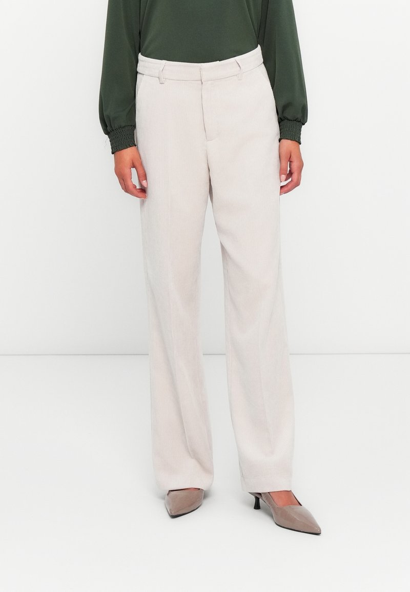 Femme portant un pantalon droit beige clair taille haute avec des poches, associé à des talons pointus taupe et un haut vert foncé à manches longues.