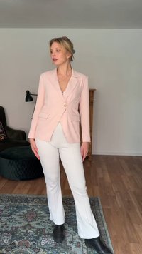 Γυναίκα στέκεται σε μοτίβο χαλί στο σαλόνι, φοράει ανοιχτό ροζ σακάκι, λευκό παντελόνι και μαύρες μπότες, κοιτάζοντας προς τα πλάγια.