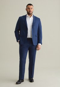 Completo blu navy realizzato in tessuto texturizzato, caratterizzato da un blazer a un bottone e pantaloni con frontale piatto, abbinato a una camicia bianca e scarpe nere.