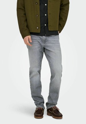 Homme portant un jean droit gris délavé, des chaussures en cuir marron foncé avec des lacets roses, une chemise noire et une veste vert olive.