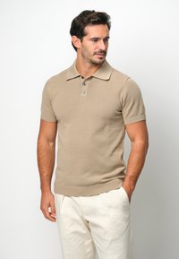 Beige kortärmad polo med en strukturerad stickad mönster, tvåknapplåsning och fast nederkant, kombinerad med ljusa byxor.