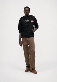 Ung man som står, klädd i svart zip-up huvtröja med röd logotyp, bruna vida byxor och bruna läderskor mot en helt vit bakgrund.