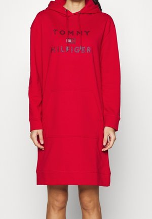 Rød langærmet hoodie-kjole med frontlomme og "Tommy Hilfiger"-logo udsmykket i blå, båret af en person, der står mod en ensfarvet baggrund.
