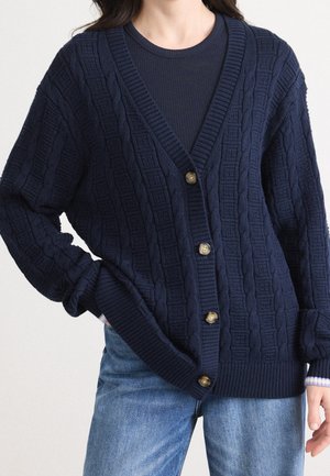 Kvinde iført en marineblå kabelstrikket cardigan med træknapper over en marineblå ribbet top og blå denimjeans, hånd i lommen.