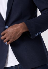 Blazer navy in tessuto texturizzato, con bottoni blu e una singola tasca. Indossato su una camicia bianca con dettagli visibili sui polsini.