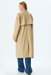 Beige trenchcoat med ett lagerdesign, med hög krage, långa ärmar och en ryggventil, kombinerad med vida denimbyxor.