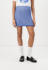 Blue and white patterned mini skirt made of textured fabric, paired with white socks and black loafers, model stands against a plain background.