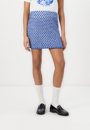 Minirock mit blau-weißen Mustern aus strukturiertem Stoff, kombiniert mit weißen Socken und schwarzen Slippern, Modell steht vor einem schlichten Hintergrund.