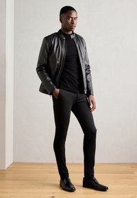 Zwarte leren jack met een hoge kraag, gecombineerd met een gestructureerde zwarte trui en slanke zwarte broek, compleet met zwarte schoenen. Achtergrond van houten vloer.
