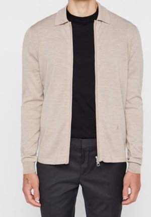 Beige vest met rits, open gedragen over een zwart shirt met ronde hals, gecombineerd met een donkerkleurige broek, gedragen door een staande persoon.