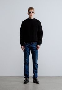 Suéter de punto acanalado negro, vaqueros de mezclilla azules y zapatos negros. El modelo lleva gafas de sol oscuras, de pie contra un fondo claro liso.