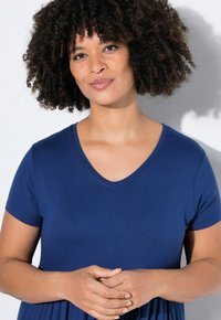 Granatowa sukienka z dekoltem w serek, krótki rękaw, z miękkiego materiału, luźny krój, z marszczonym wcięciem w talii; modelka z kręconymi włosami pozuje ze złożonymi dłońmi.