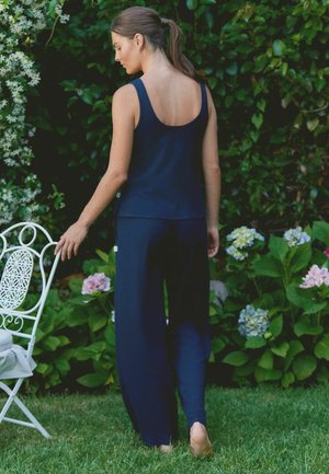 Woman in a navy sleeveless top and wide-legged pants stands barefoot on grass, holding a white metal garden chair with flowering plants in the background.