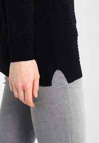 Close-up of a person’s hand resting near the hip, wearing a black textured sweater with side slit and light gray jeans.