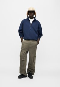 Μπλε σκούρο φούτερ με φερμουάρ, μπεζ καπέλο κουβάς με προστατευτικά για τα αυτιά, ελιάς παντελόνι cargo με τσέπες, μαύρα αθλητικά παπούτσια, στέκεται μπροστά σε λευκό φόντο.